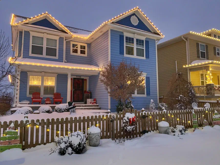 Blue two-story home with warm white roofline lights and a snow-covered yard