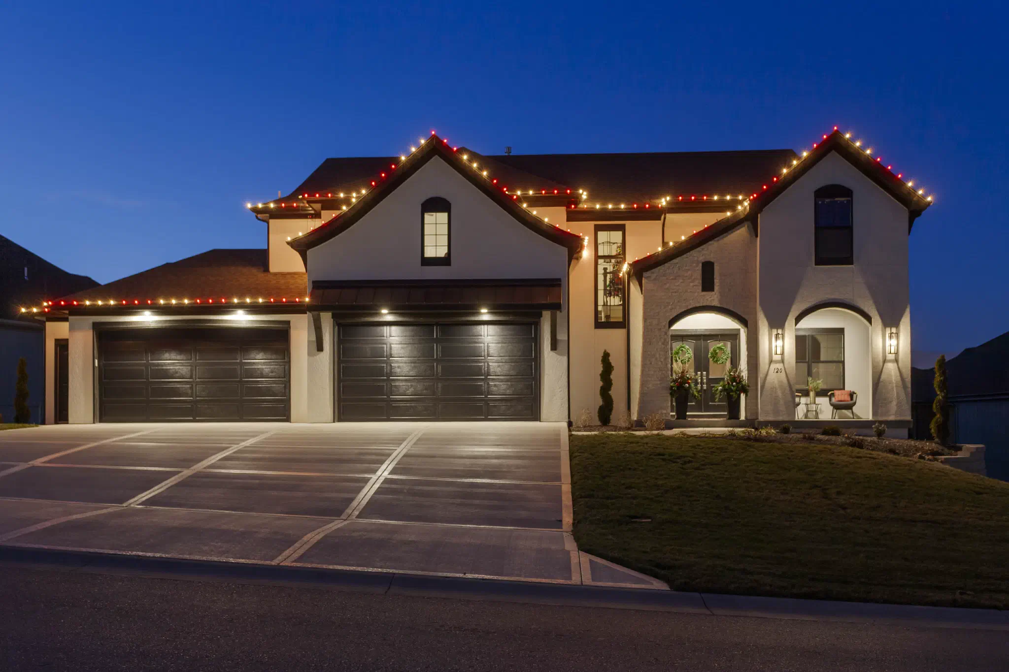 Stucco home with three-car garage outlined in red and warm white holiday lights at dusk